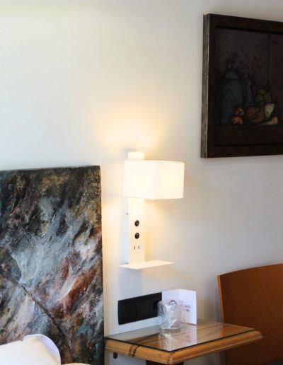 Un dormitorio moderno y luminoso con una pared blanca. A la izquierda, se ve parcialmente una cabecera de cama con un cuadro abstracto en tonos azules, marrones y ocres.