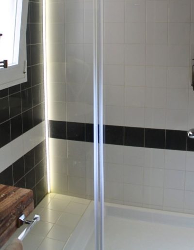 Un baño moderno con una ducha de mampara de cristal transparente. Las paredes de la ducha están revestidas con azulejos blancos y una banda horizontal de azulejos negros. Dentro de la ducha, se ven dispensadores de jabón y una barra de toalla. A la izquierda de la ducha, hay un lavabo de cuenco redondo sobre un mueble rústico de madera, y un espejo de aumento montado en la pared. Una ventana con marco blanco se ve en la parte superior izquierda.