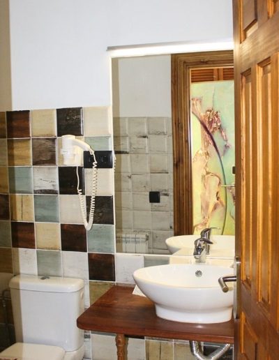 Un cuarto de baño con paredes cubiertas de azulejos patchwork en tonos tierra, grises, verdes y blancos, con un estilo rústico. A la izquierda, se ve la cisterna de un inodoro blanco. En el centro, un lavabo blanco ovalado con grifo cromado se asienta sobre una encimera de madera.