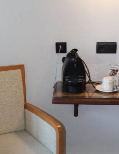 Una silla con marco de madera clara y tapicería beige en primer plano a la izquierda. A la derecha, una mesita flotante de madera con una superficie de cristal. Sobre ella, hay una cafetera negra, dos tazas de café blancas con sus platillos y una caja pequeña. En la pared blanca, detrás de la mesa, se ven varios enchufes negros.