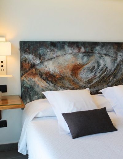 Una cama grande con ropa de cama blanca y dos cojines decorativos de color gris oscuro. La cabecera de la cama es una obra de arte abstracta y texturizada en tonos azules, marrones y blancos, que sugiere un patrón en espiral o movimiento.