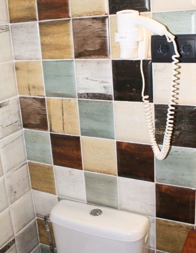 Una pared de baño cubierta con azulejos patchwork en tonos marrones, grises, blancos y verdosos, con un aspecto envejecido o rústico. En la parte superior derecha, hay un secador de pelo blanco montado en la pared, con su cable en espiral.