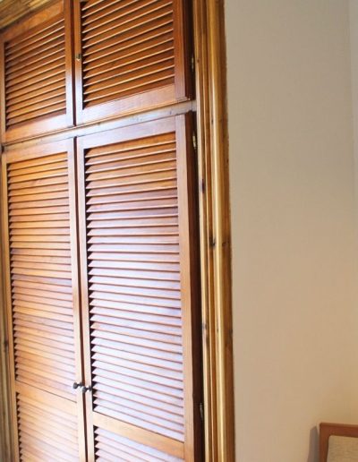 Un armario alto y estrecho de madera con acabado en tono miel. Presenta cuatro puertas de persiana, dos en la parte superior más pequeñas y dos más grandes debajo, todas con tiradores oscuros. El marco del armario también es de madera. El armario está empotrado en una pared de color claro.