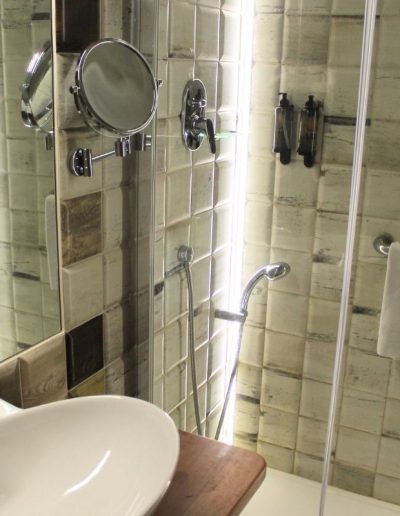 Una sección de un cuarto de baño moderno con un lavabo blanco ovalado y un grifo cromado en primer plano, sobre una encimera de madera. Al fondo, se observa una mampara de ducha de cristal transparente, detrás de la cual hay azulejos texturizados en tonos neutros o ligeramente envejecidos.