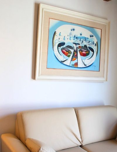 Un sofá de color crema claro con un cojín a juego en el lado derecho. Sobre el sofá, colgado en una pared blanca, hay un cuadro con un marco grueso de color crema y un paspartú a juego. La pintura es abstracta con un fondo azul claro, representando lo que parecen ser cestas de frutas en primer plano y un paisaje con palmeras en el fondo.