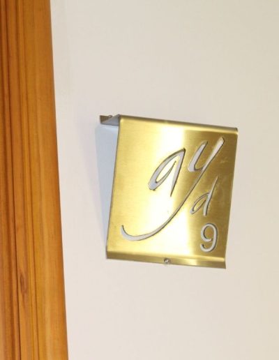 Una placa rectangular de metal dorado con un diseño calado del logo 'ayd' y el número '9' en la parte inferior. La placa está fijada a una pared clara, posiblemente de un hotel o establecimiento, y se ve parte de un marco de puerta de madera clara a la izquierda.
