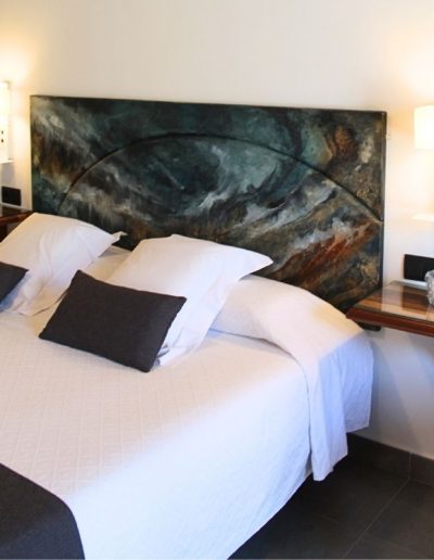 Una cama de matrimonio con sábanas blancas, almohadas blancas y dos cojines negros. El cabecero es una pieza grande de arte abstracto en tonos oscuros de azul, verde y marrón rojizo. A cada lado de la cama, hay mesitas de noche flotantes de madera con lámparas de pared blancas y vasos de agua. Un letrero redondo de prohibido fumar se ve en la mesita de noche derecha.
