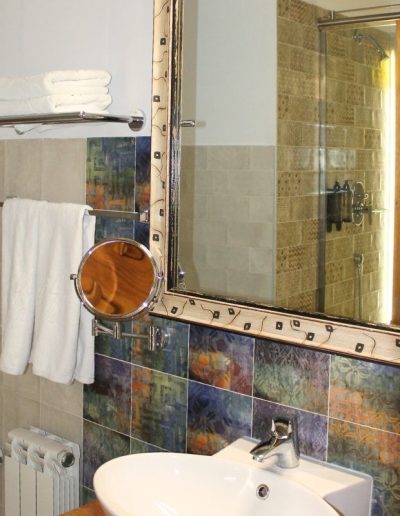 Un baño con un estilo moderno y colorido. La pared está decorada con azulejos patchwork en tonos azules, verdes, ocres y grises en la parte inferior, y azulejos lisos en la parte superior. Un gran espejo enmarcado con un diseño abstracto en negro y dorado domina el centro, reflejando una zona de ducha con azulejos texturizados.