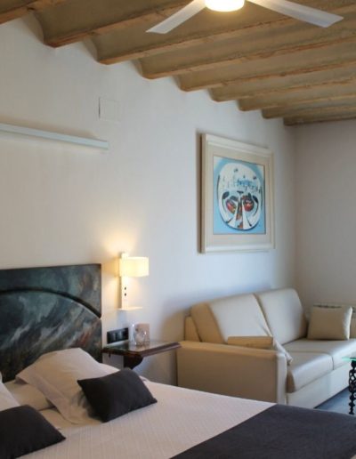 Una habitación espaciosa con una cama a la izquierda, que tiene un cabecero abstracto en tonos oscuros y dos lámparas de pared blancas a cada lado. En el centro de la habitación, hay un sofá de cuero crema con un cojín claro y una mesa de centro de cristal y metal. Un cuadro en tonos azules y blancos cuelga sobre el sofá. El techo muestra vigas de madera expuestas y un ventilador de techo blanco con luz central.
