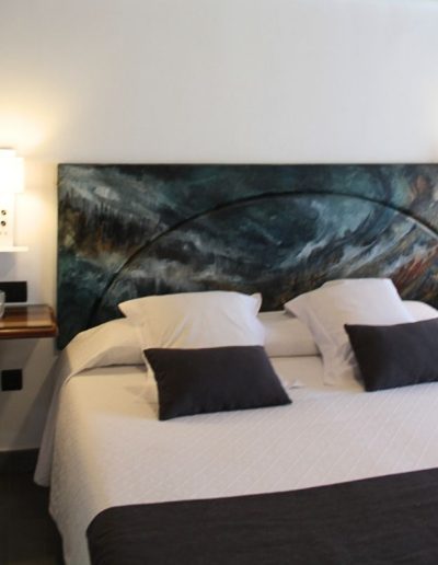 Una cama de matrimonio con sábanas blancas, almohadas blancas y cojines decorativos negros. El cabecero es una pieza de arte abstracta en tonos verdes oscuros, azules y ocres, con una forma curva prominente. A cada lado de la cama, hay mesitas de noche flotantes de madera oscura con lámparas de pared blancas que emiten una luz cálida.