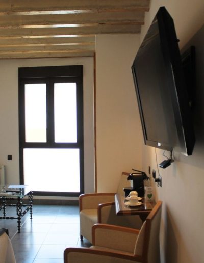 Una sala de estar o habitación de hotel con una televisión de pantalla plana montada en la pared. Debajo de la televisión hay una pequeña mesa flotante de madera con una cafetera, tazas de café y un vaso. Delante de la mesa hay dos sillas de tela de color claro. Al fondo, se ve un sofá de color claro, una mesa de centro de cristal y una ventana alta y estrecha con marco oscuro, que deja entrar mucha luz. El techo tiene vigas de madera vistas.