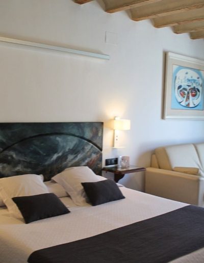 Una cama de matrimonio con sábanas blancas, almohadas blancas y dos cojines negros. El cabecero es una pieza de arte abstracto en tonos oscuros de verde y azul. A la derecha de la cama, una mesita de noche flotante de madera tiene una lámpara de pared blanca y un vaso. Al fondo de la habitación, se ve un sofá de color crema y, sobre él, un cuadro con un marco blanco y una pintura abstracta con formas circulares en tonos azules y blancos. El techo de la habitación tiene vigas de madera vistas.