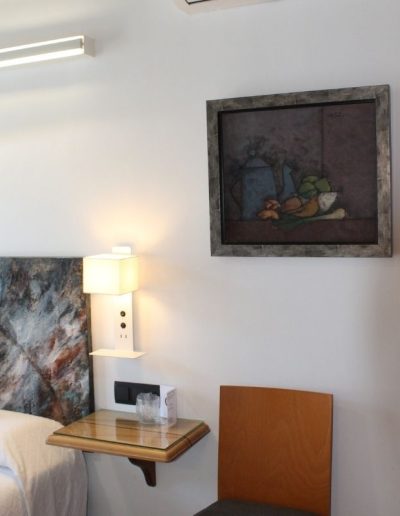 Una sección de un dormitorio moderno con una pared blanca. Se ve parte de una cabecera de cama con un diseño abstracto y texturizado en tonos oscuros y ocres. Junto a la cama, hay una mesita de noche flotante con superficie de cristal y una lámpara de aplique blanca de diseño contemporáneo que emite luz hacia arriba y hacia abajo.