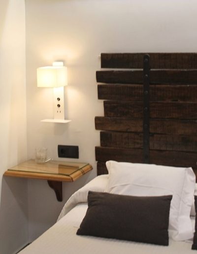 Una cama de matrimonio con sábanas claras, almohadas blancas y cojines oscuros. El cabecero es de madera rústica oscura, con listones horizontales. A la izquierda de la cama, una mesita de noche flotante de madera con superficie de cristal tiene una lámpara de pared blanca que emite luz cálida y un vaso con agua.
