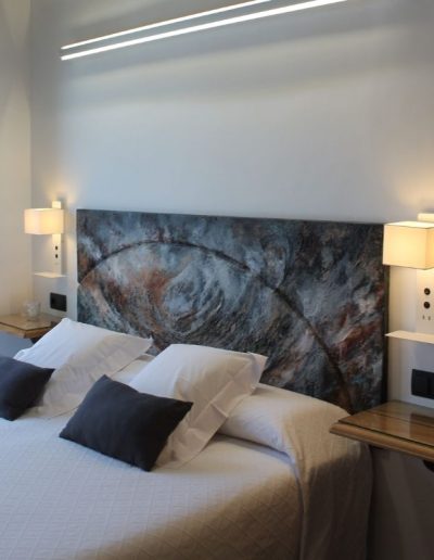 Una cama grande con ropa de cama blanca y cojines grises oscuros. La cabecera es una obra de arte abstracta con textura, en tonos oscuros como azul, gris y ocre. A ambos lados de la cama, hay mesitas de noche flotantes con superficies de madera y cristal, y lámparas de aplique blancas modernas que emiten una luz cálida.