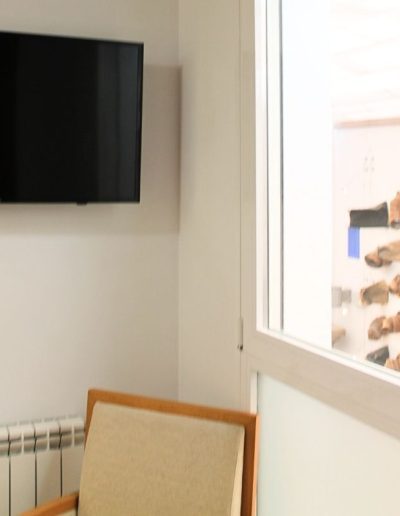 Una esquina de una habitación con una televisión de pantalla plana montada en una pared blanca a la izquierda. Debajo de la televisión, se ve parte de un radiador blanco y, en primer plano, el brazo y parte del asiento de una silla con marco de madera clara y tapicería beige. A la derecha, una ventana con marco blanco permite ver el interior de otra habitación donde hay una instalación artística en la pared con múltiples objetos de madera o cerámica de tonos tierra.