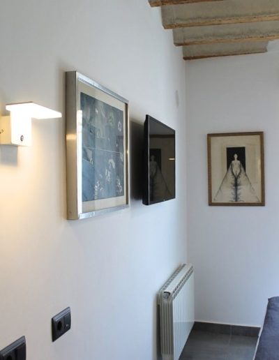 Una pared blanca con techo de vigas de madera expuestas en la parte superior derecha. En la pared, hay una lámpara de aplique blanca de diseño moderno. Debajo, se aprecian varios enchufes e interruptores oscuros.