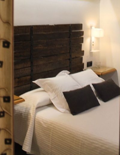 El reflejo de una cama de matrimonio en un espejo con marco ornamentado y oscuro. La cama tiene sábanas blancas con un patrón sutil, almohadas blancas y cojines negros. El cabecero de la cama es de madera oscura con listones horizontales.