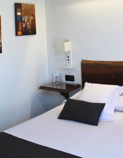 "Una cama con ropa de cama blanca y cojines decorativos de color gris oscuro. La cabecera es una pieza de madera oscura y maciza con el borde superior natural e irregular. A la izquierda, en la pared blanca, cuelgan dos cuadros abstractos de formato vertical.