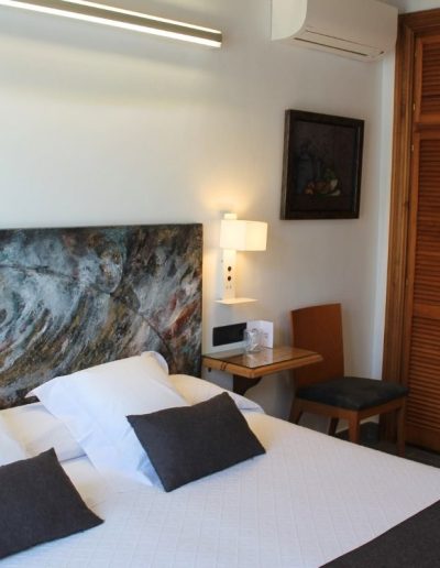 Un dormitorio moderno y luminoso con una cama grande vestida con ropa de cama blanca y cojines grises. La cabecera de la cama es un cuadro abstracto con tonos azules, marrones y ocres. A la derecha de la cama, hay una mesita de noche con superficie de cristal y una lámpara de aplique blanca contemporánea que ilumina la zona.