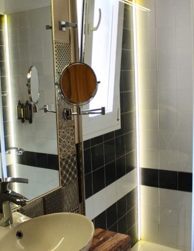 Un baño moderno con paredes de azulejos blancos y una banda horizontal de azulejos negros. En primer plano, se ve un lavabo de cuenco blanco con un grifo cromado, situado sobre un mueble de madera rústica. Un gran espejo se extiende por encima del lavabo, reflejando una ventana abierta y un espejo de aumento con marco de madera.