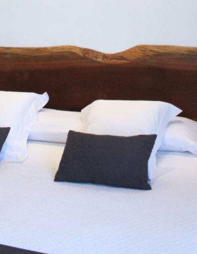 Una cama con ropa de cama blanca y dos almohadas blancas con dos cojines decorativos de color gris oscuro. La cabecera de la cama es una gran pieza de madera oscura y maciza, con el borde superior natural e irregular, lo que le da un aspecto rústico y orgánico.