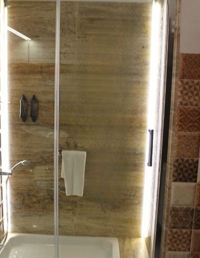 Una cabina de ducha con puertas de cristal transparentes y paredes revestidas con azulejos de imitación mármol en tonos marrones y beige. En la pared izquierda de la ducha, se ve una alcachofa de ducha cromada y botellas de amenidades en un dispensador doble. Una toalla blanca cuelga de un riel dentro de la ducha. A la derecha de la ducha, la pared está cubierta con azulejos decorativos de patrón geométrico en tonos marrones y blancos.